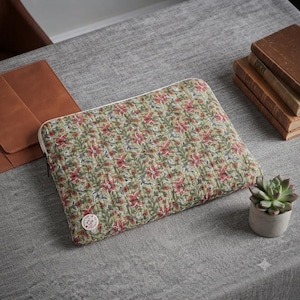 Puede incluir: Una funda rectangular acolchada para portátil hecha de tela verde oliva con un patrón floral repetitivo en tonos rojos, rosas y blancos. La funda cuenta con un cierre de cremallera y una pequeña etiqueta de marca redonda y blanca.