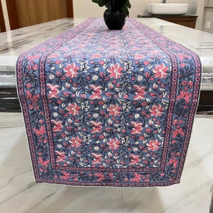 May include: A blue floral table runner with a pink and white flower pattern. The runner has a dark blue border with a pink geometric design. The runner is placed on a marble table.