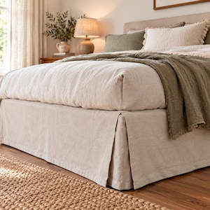 May include: A beige linen bed valance with a tailored pleated design, covering the base of a bed dressed in a light beige duvet, olive green throw blanket, and textured pillows, creating a neutral layered bedding arrangement.