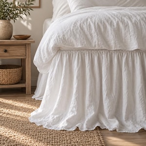 Può includere: Una balza per letto bianca con un design arricciato e a volant che pende dalla base del letto. Il tessuto ha un aspetto testurizzato e leggermente stropicciato, copre la parte inferiore della struttura del letto e scende fino al pavimento.
