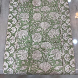 May include: A rectangular, green and white floral patterned table runner. The runner features a repeating pattern of large white flowers and foliage on a green background. The edges have a scalloped design in white and green.