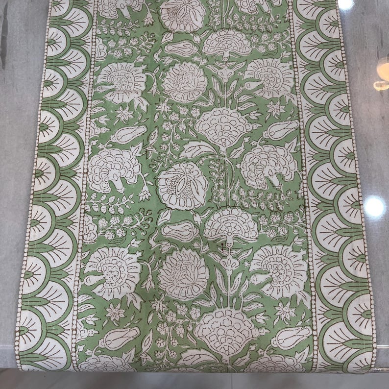 May include: A rectangular, green and white floral patterned table runner. The runner features a repeating pattern of large white flowers and foliage on a green background. The edges have a scalloped design in white and green.