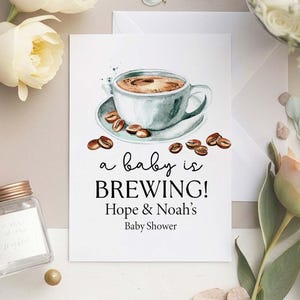 May include: A rectangular white card featuring a watercolor illustration of a light blue coffee cup on a saucer surrounded by scattered coffee beans. The text reads: a baby is BREWING! Hope & Noah's Baby Shower in black script and serif fonts.