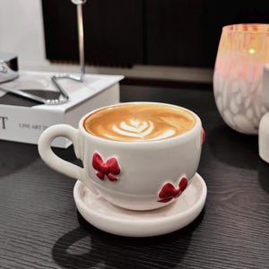Könnte beinhalten: Ein weißes Keramik-Tassen- und Untertassen-Set mit roten Schleifenakzenten. Die Tasse ist mit einem Latte gefüllt, der ein dekoratives Schaummuster aufweist. Das Set steht auf einer dunklen Oberfläche, was auf ein Café oder eine häusliche Umgebung hindeutet.