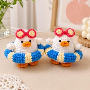 Puede incluir: Dos patitos de ganchillo hechos a mano, cada uno con gafas de sol con borde rojo y un flotador a rayas azules y amarillas. Los patitos tienen cuerpos blancos, picos y patas naranjas y ojos negros pequeños. El fondo es de color neutro.