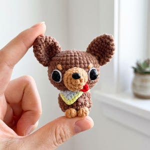 May include: A small brown crocheted chihuahua figurine with large black plastic eyes, a tiny black nose, and a small red tongue sticking out. The dog wears a light yellow and blue triangular bandana around its neck and has tan paws.