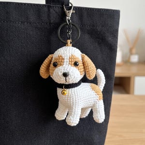 Peut inclure: Un porte-clés chien en crochet représentant un chiot blanc et beige avec des yeux noirs, un collier noir et une petite clochette dorée. Le chien en peluche est fixé à un anneau et un mousqueton en métal noir sur un sac fourre-tout noir.