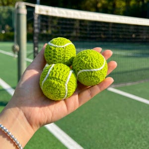Könnte beinhalten: Drei kleine, runde, lindgrüne gehäkelte Kugeln mit weißen Garnverzierungen, die die geschwungenen Linien eines Tennisballs nachahmen. Die strukturierten, handgefertigten Garnobjekte sind in einer Gruppe angeordnet und zeigen das komplexe Maschenbild.