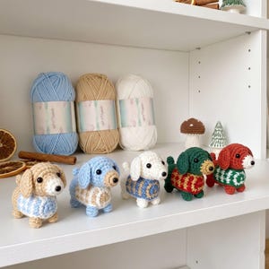 Può includere: Cinque piccole statuette di bassotto all'uncinetto disposte in fila, ognuna con un maglione di colore diverso. Dietro ci sono tre gomitoli di lana in azzurro, beige e bianco, insieme a piccoli oggetti decorativi come un fungo e alberi.