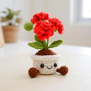 May include: A crocheted flower pot with a smiling face, holding a red flower with green leaves and a bud. The pot is white with brown soil and two brown crocheted feet.
