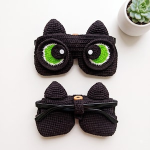 May include: Two black crocheted cat-shaped eyeglass cases. The top case features large, bright green eyes with white pupils. The bottom case shows the back side with a small wooden button closure securing a pair of black-framed eyeglasses inside.