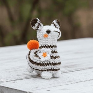 May include: A handmade crochet llama figurine. The white llama has brown and white striped accents, a small orange pom-pom, and a flower with an orange center. The llama is standing on a white wooden surface.
