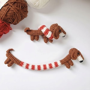 May include: Two crocheted dachshund figures made of brown yarn with white and red striped midsections. Each dog features small black eyes, a white muzzle, and a black nose. The figures are crafted with a textured stitch pattern and appear soft.