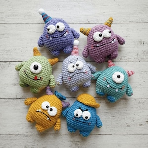 Puede incluir: Colección de siete juguetes de monstruos de ganchillo coloridos. Cada monstruo tiene un color, una expresión facial y pequeños cuernos o gorros de fiesta únicos. Los juguetes están dispuestos sobre una superficie de madera clara.