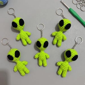 Könnte beinhalten: Fünf limegrüne gehäkelte Alien-Schlüsselanhänger mit großen schwarzen ovalen Augen. Jede Figur hat einen kleinen Kopf, einen schlanken Körper und kurze Gliedmaßen, die mit einer kurzen Kette an einem silberfarbenen Metallring befestigt sind.