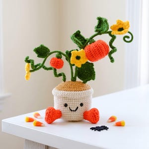 May include: A crocheted pumpkin plant in a beige pot with a smiling face and orange feet. The plant features green vines, dark green leaves, bright yellow flowers, and two orange pumpkins. Several candy corn pieces and a small black spider are nearby.