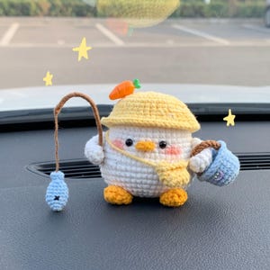 Pode incluir: Uma figura de patinho de crochê branca usando um chapéu amarelo com um detalhe de cenoura laranja. O patinho segura uma vara de pescar marrom com um peixe azul claro e carrega um pequeno balde azul claro com alça marrom ao lado.
