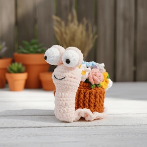 Puede incluir: Un caracol de ganchillo hecho a mano con ojos grandes, blancos y negros, un cuerpo rosa claro y una concha marrón llena de flores de ganchillo coloridas. El caracol está sobre una superficie de madera blanca.