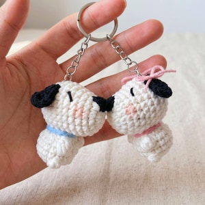 May include: A crochet keychain featuring two small white puppies with black ears and noses. One puppy wears a light blue collar, while the other has a pink collar and a small pink bow on its head. Both figures are attached to a silver metal ring.