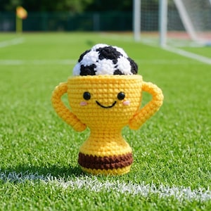 May include: A yellow crocheted trophy with a smiling face and a black and white soccer ball inside. The trophy has two handles and a brown stripe at the base, set against a green field.