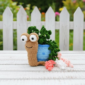 Puede incluir: Una maceta de caracol de ganchillo hecha a mano con un cuerpo marrón claro, ojos grandes y saltones y una cara sonriente. La maceta azul contiene plantas de ganchillo verdes, con flores de ganchillo rosas y blancas en la base. El caracol está contra una valla blanca.