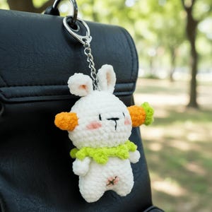 May include: A white crochet bunny keychain with orange accents on the ears and a green collar. The bunny has a black face and pink cheeks. The keychain is attached to a silver metal clasp and chain, hanging from a black bag.