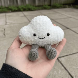 May include: A white plush cloud with a smiling face and gray crocheted legs. The cloud is soft and fluffy, with black eyes and a curved mouth. A cute, whimsical toy.