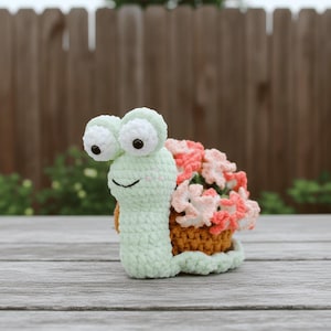 Puede incluir: Un caracol de ganchillo hecho a mano con grandes ojos saltones y una cara sonriente. La concha del caracol está llena de flores rosas y melocotón. El caracol es de color verde claro con una concha marrón.