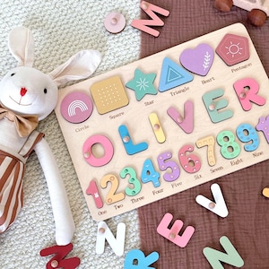 May include: A wooden puzzle board featuring the name OLIVER, numbers 1 through 9, and various shapes including a circle, square, star, triangle, heart, and pentagon. Each piece has a small wooden knob. Loose letters are scattered nearby.