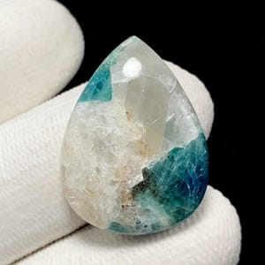 May include: A pear-shaped gemstone featuring a faceted top section and a rough, textured lower portion. The stone displays a colour gradient transitioning from translucent white and clear crystal at the top to deep teal and blue hues at the base.