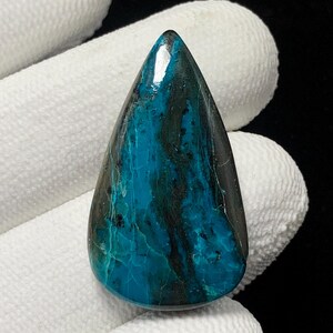 May include: A teardrop-shaped gemstone with a polished surface. The stone features a blend of teal and black hues, with a glossy finish. The gemstone is held against a white background.
