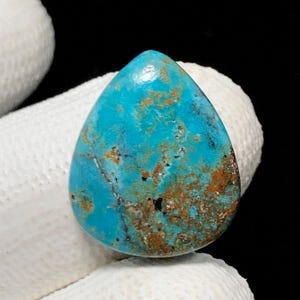 Puede incluir: Una gema de turquesa pulida en forma de lágrima. La piedra presenta un tono azul vibrante con patrones de matriz marrón y negra. La gema es sostenida por una mano enguantada, mostrando su belleza natural.