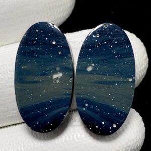 Puede incluir: Dos piedras preciosas ovaladas en azul oscuro y gris, que recuerdan a un cielo estrellado. Las piedras presentan motas blancas y una superficie pulida, ideales para la fabricación de joyas o coleccionar.
