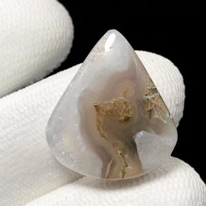 Könnte beinhalten: Ein polierter, dreieckiger Edelstein mit einer Mischung aus weißen, braunen und beigen Farben. Der Stein hat eine durchscheinende Qualität mit wirbelnden Mustern und einer glatten, glänzenden Oberfläche. Der Edelstein wird vor einem weißen Stoffhintergrund gehalten.