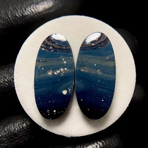May include: Two oval-shaped cabochons with a dark blue, swirling pattern reminiscent of a starry night sky. The cabochons have white and silver accents, and are set against a white background.