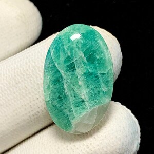 May include: An oval-shaped cabochon gemstone featuring a vibrant teal and seafoam green color palette. The stone displays a complex internal structure with visible crystalline fractures, white inclusions, and a polished, smooth surface finish.