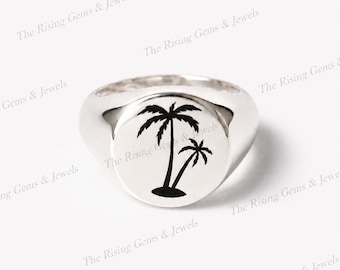 May include: A polished silver signet ring featuring a flat, circular face engraved with a black graphic of two palm trees. The ring has a smooth, rounded band and a reflective metallic finish, showcasing a clean design with a tropical motif.