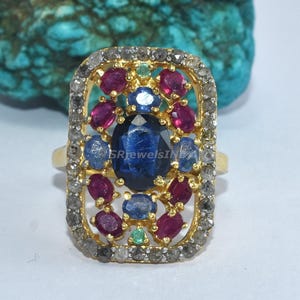 Könnte beinhalten: Ein goldener Ring mit rechteckiger Fassung, der einen großen ovalen blauen Edelstein in der Mitte aufweist, umgeben von kleineren roten und blauen Steinen. Die Fassung des Rings wird von einer Bordüre aus kleinen, grauen Steinen umrahmt. Der Ring ist vor einem türkisfarbenen Hintergrund platziert.