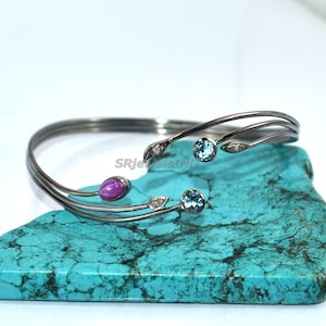 May include: A silver-toned cuff bracelet with a delicate, flowing design. The bracelet features a magenta-colored gemstone and two light blue gemstones, along with small clear accents. The bracelet is displayed on a turquoise stone.