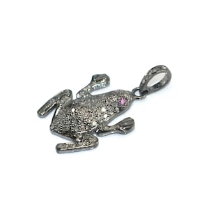 Può includere: Un ciondolo a forma di rana grigio scuro con un piccolo anello per una catena. La rana è ricoperta di piccole pietre scintillanti e ha una singola pietra rosa per un occhio. Il ciondolo è su sfondo bianco.