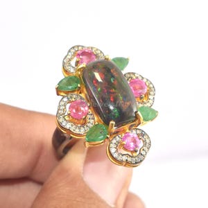 Op de afbeelding: Een goudkleurige ring met een grote, rechthoekige zwarte opaal. De opaal is omgeven door roze en groene edelstenen en kleine diamanten. De ring heeft een bloemmotief, waarbij de edelstenen zijn gerangschikt als bloemen.