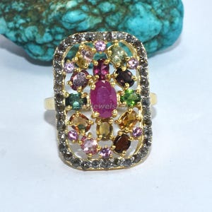 May include: A gold ring with a rectangular face, encrusted with a variety of colorful gemstones. The central stone is a large, oval ruby, surrounded by smaller gems in shades of pink, green, yellow, and brown. The ring's setting is detailed with small, dark stones.