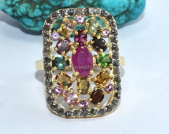 May include: A gold ring featuring a rectangular face adorned with an array of colourful gemstones. A large, oval ruby is at the centre, encircled by smaller gems in pink, green, yellow, and brown. The ring's setting is detailed with small, dark stones.