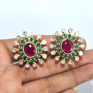Op de afbeelding: Een paar goudkleurige oorstekers. Elke oorbel heeft een grote, ovale, dieprode edelsteen in het midden, omringd door kleinere groene en witte edelstenen. De oorbellen hebben een sunburst-ontwerp, met de edelstenen in een cirkelvormig patroon.