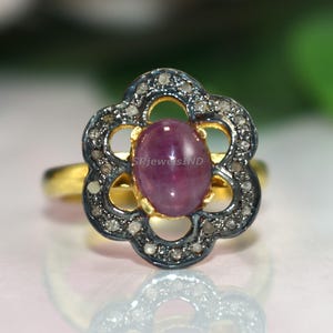 Peut inclure: Une bague en or avec un sertissage en forme de fleur. Le sertissage a une bordure gris foncé avec de petites pierres claires et une pierre centrale ovale, rose rougeâtre. La bague est sur une surface réfléchissante.