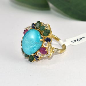 May include: A gold ring featuring a large, oval turquoise stone as the centerpiece. The ring is adorned with smaller gemstones in shades of green, red, and blue, and is set with small, clear accent stones. The ring has a classic, ornate design.