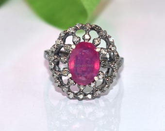 May include: A silver ring featuring a large, oval magenta gemstone. The gemstone is encircled by a detailed, openwork design with small, clear accent stones. The ring is set against a white backdrop.