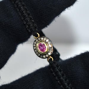 May include: A black macrame bracelet featuring a gold-toned oval centerpiece. The centerpiece is adorned with a central pink gemstone, surrounded by small white gemstones. The bracelet is held by a black gloved hand.