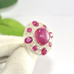 Può includere: Un anello d'argento con un grande rubino ovale al centro, circondato da rubini ovali più piccoli e un alone di minuscole pietre scintillanti. Il design dell'anello è circolare e ornato, con una finitura lucida e brillante.