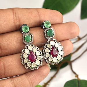 May include: A pair of dangle earrings featuring emerald and ruby gemstones. Each earring has a top section with two square emeralds, a middle section with small white stones, and a bottom section with a ruby center surrounded by white stones.